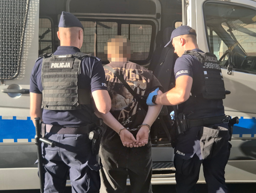 Dwóch policjantów prowadzi skutego kajdankami mężczyznę do otwartego radiowozu.