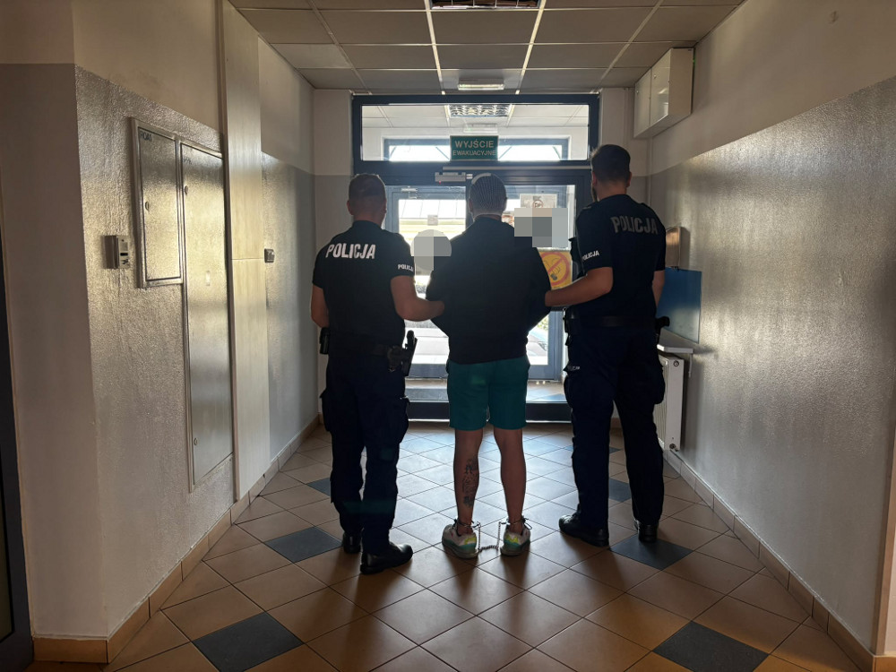 Mężczyzna w krótkich spodniach prowadzony przez dwóch policjantów w korytarzu budynku.