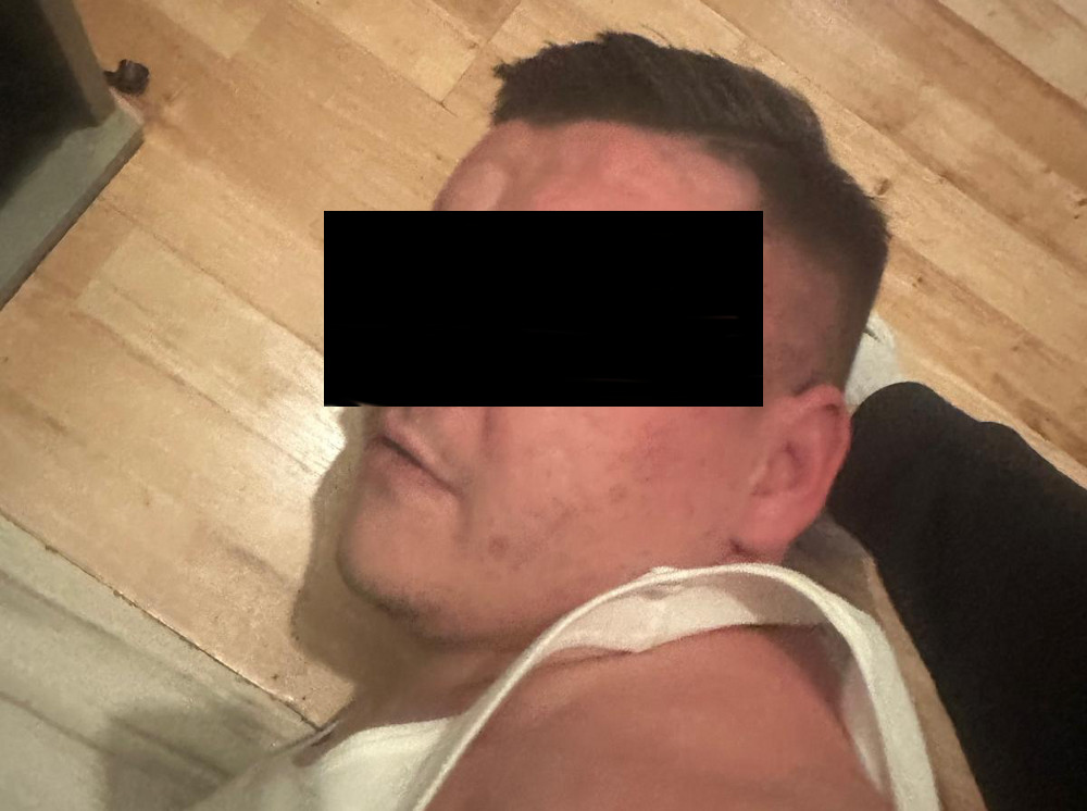Mężczyzna leży na podłodze w białym podkoszulku, z widocznym wyrazem twarzy, na tle drewnianej podłogi.