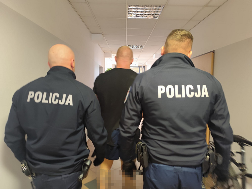 Trzech policjantów w mundurach prowadzi mężczyznę w kierunku wyjścia z budynku.