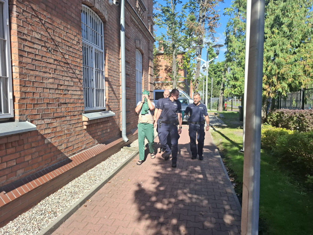 Trzech policjantów eskortuje mężczyznę w zielonym ubraniu, zasłaniającego twarz, wzdłuż chodnika przy budynku z cegły.
