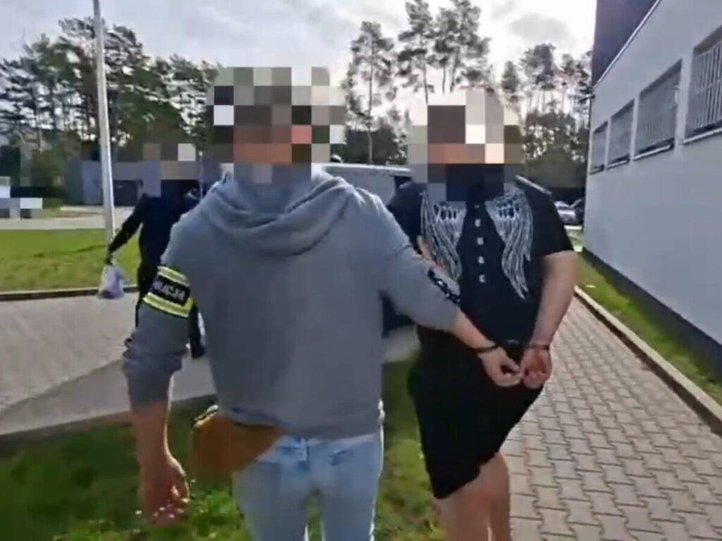 Mężczyzna w szarej bluzie prowadzi osobę w kajdankach na terenie zewnętrznym, obok budynku.
