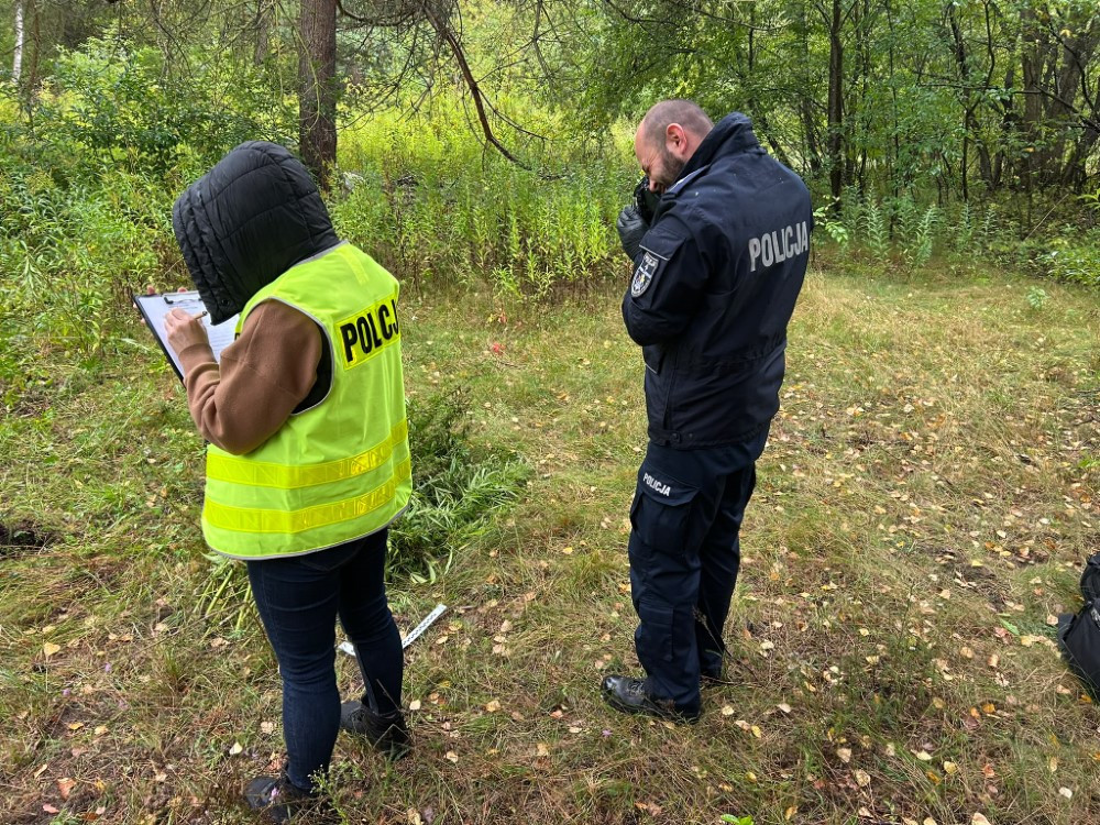 Funkcjonariusz policji w żółtej kamizelce notuje informacje, podczas gdy drugi policjant rozmawia przez telefon w lesie.