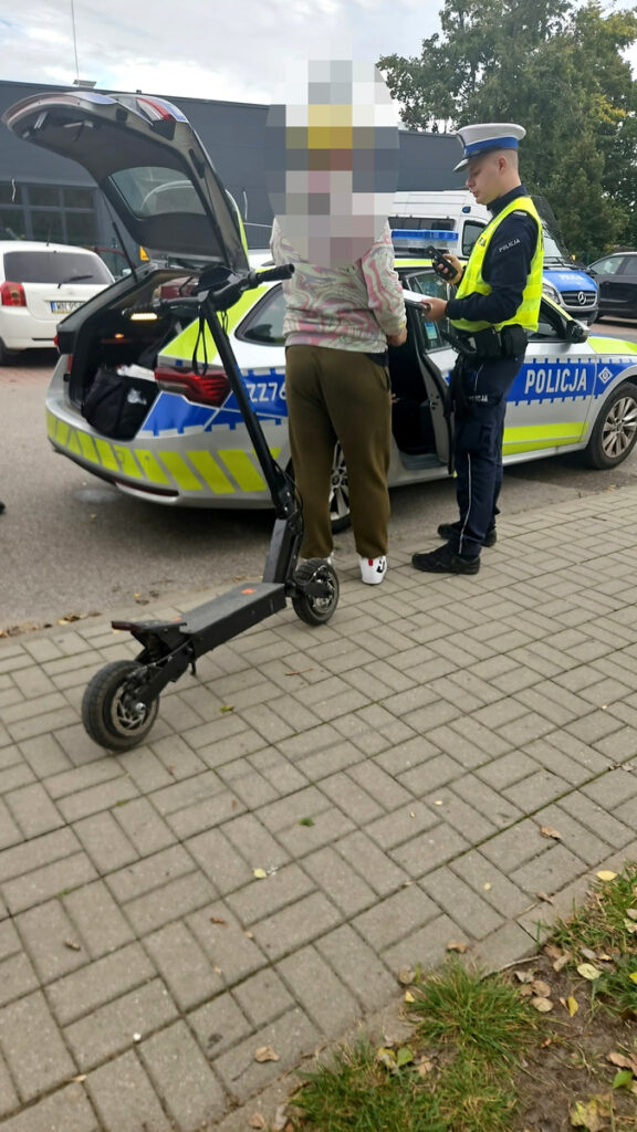 Mężczyzna rozmawia z policjantem obok radiowozu, podczas gdy skuter elektryczny stoi na chodniku.