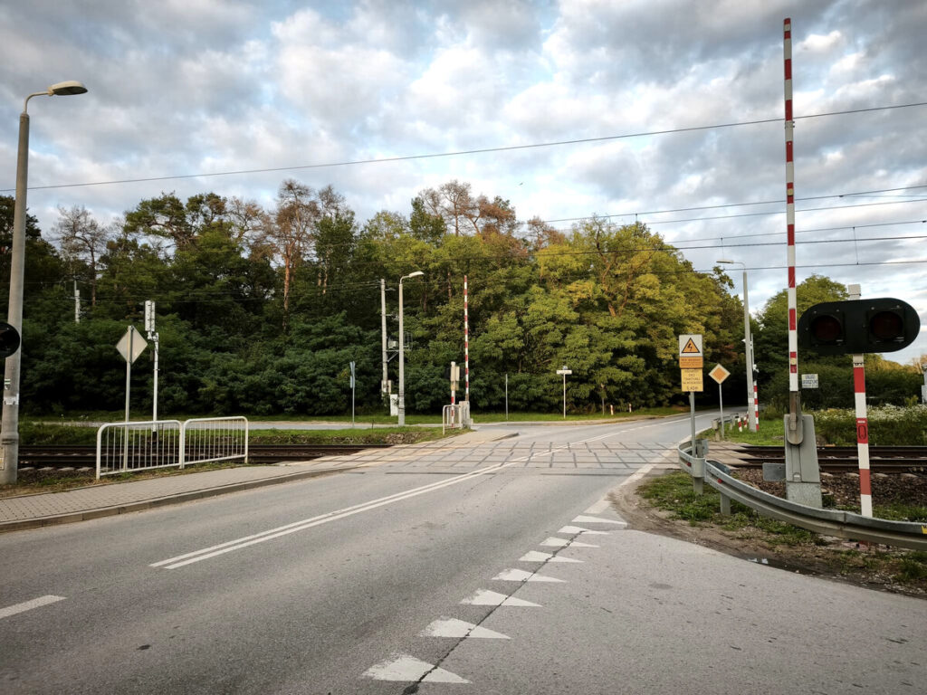 Zamknięte przejście kolejowe z sygnalizacją świetlną, otoczone zielenią drzew i chmurami na niebie.