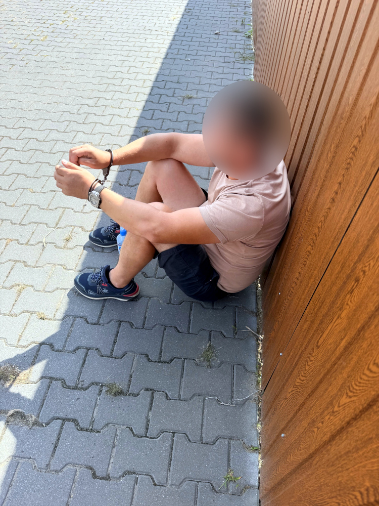 Mężczyzna w krótkich spodniach siedzi na chodniku, opierając się plecami o drewnianą ścianę.