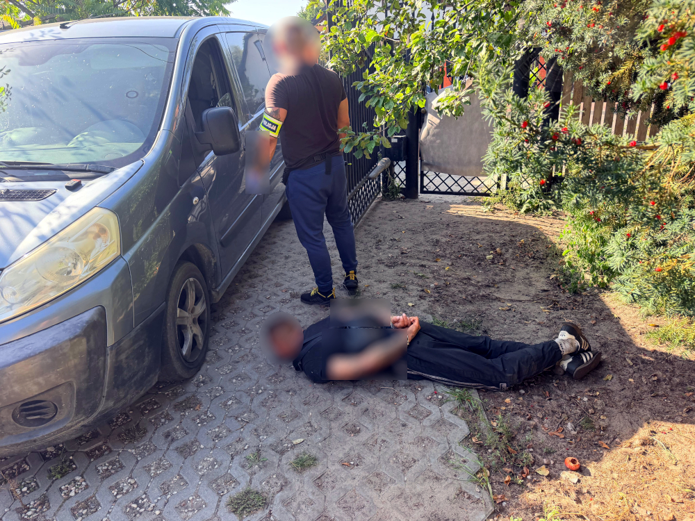 Mężczyzna leży na ziemi obok szarego furgonu, a policjant z oznakowaniem na rękawie sprawdza sytuację w ogrodzie.