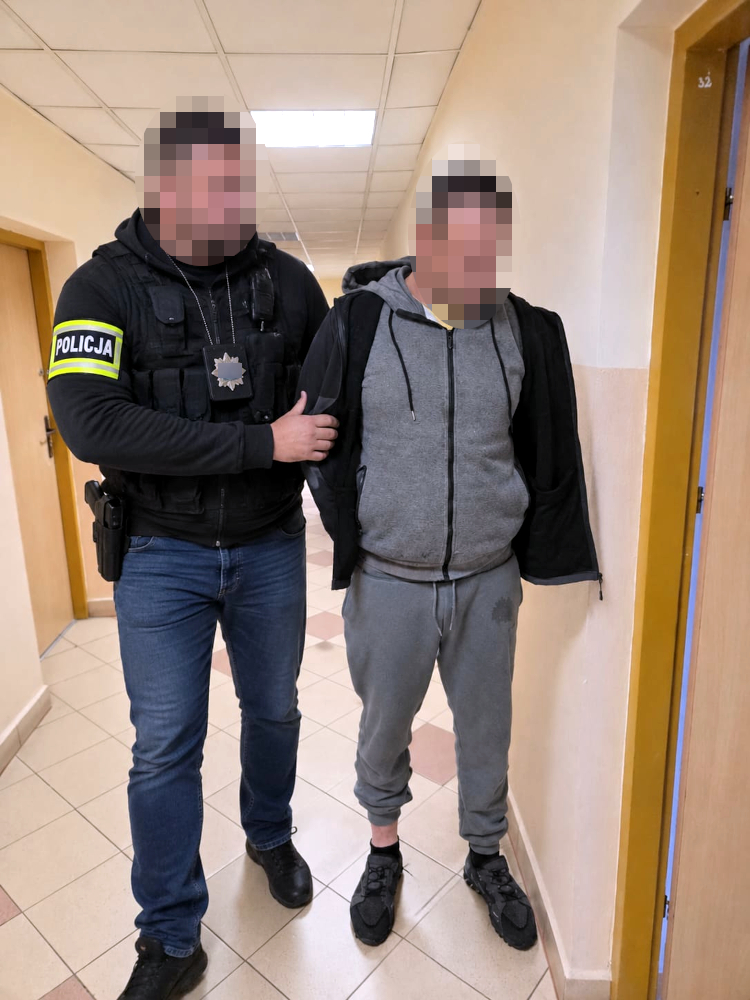 Policjant w mundurze prowadzi zatrzymanego mężczyznę w szarej odzieży przez korytarz budynku.