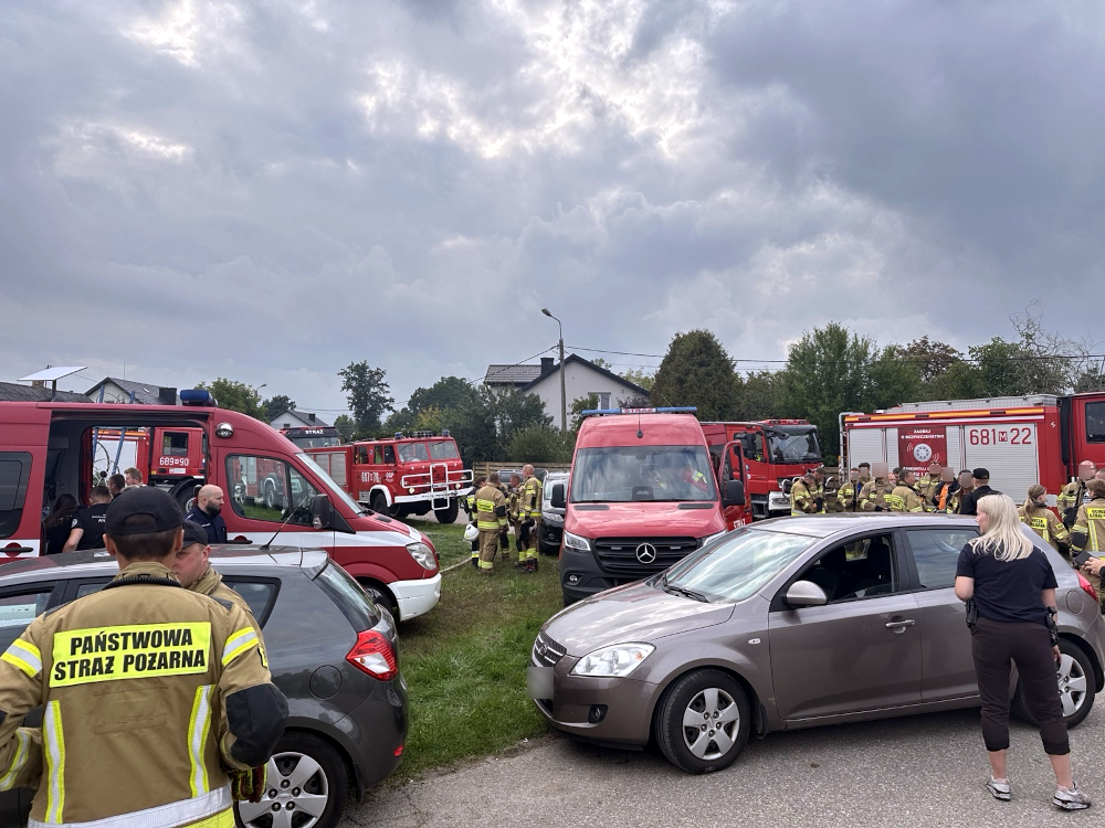 Grupa strażaków i ratowników medycznych zbiera się przy zaparkowanych wozach strażackich na terenie zdarzenia.