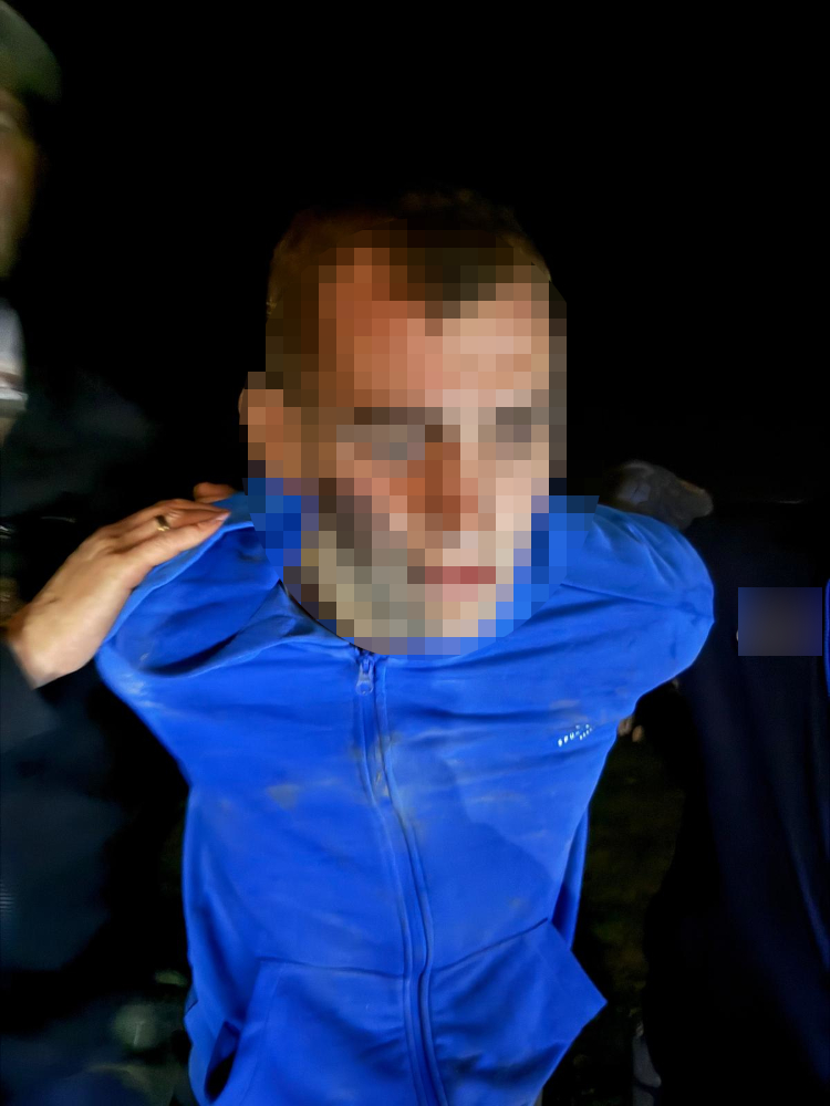 Mężczyzna w niebieskiej bluzie trzymany przez dwóch innych mężczyzn w ciemnym otoczeniu nocnym.