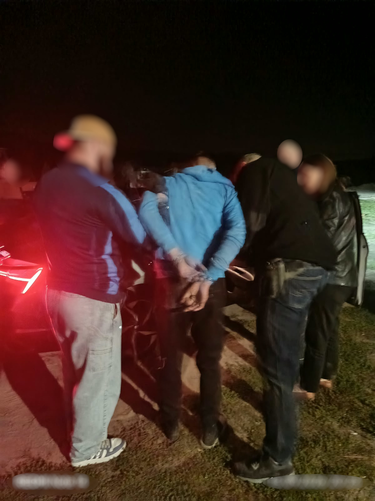 Grupa osób otacza mężczyznę w kajdankach, stojąc obok samochodu w ciemnej scenerii nocnej.