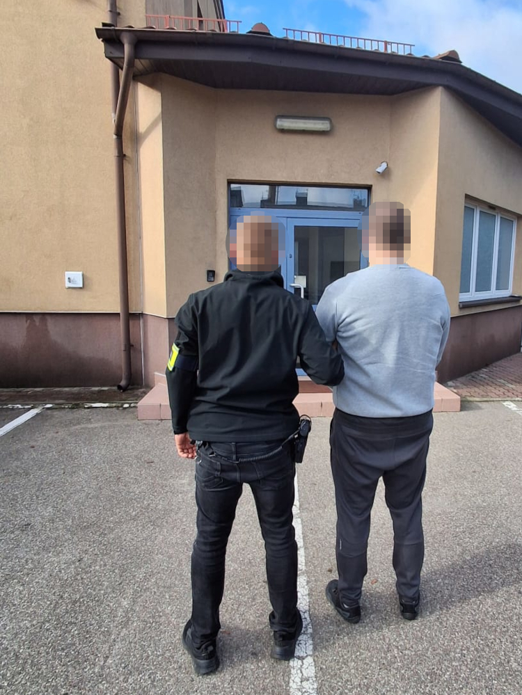 Funkcjonariusz prowadzi mężczyznę w szarej bluzie w stronę wejścia do budynku; twarze obu są zamazane.