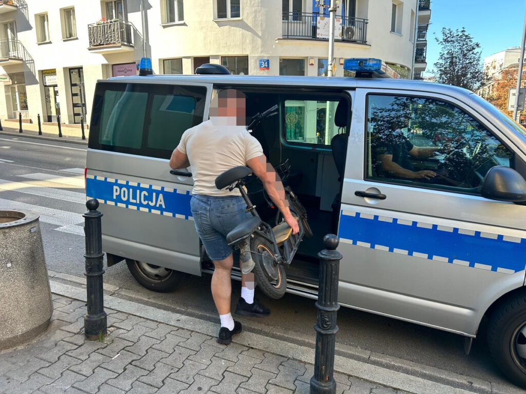Mężczyzna w krótkich spodniach wkłada rower do policyjnego samochodu na ulicy.