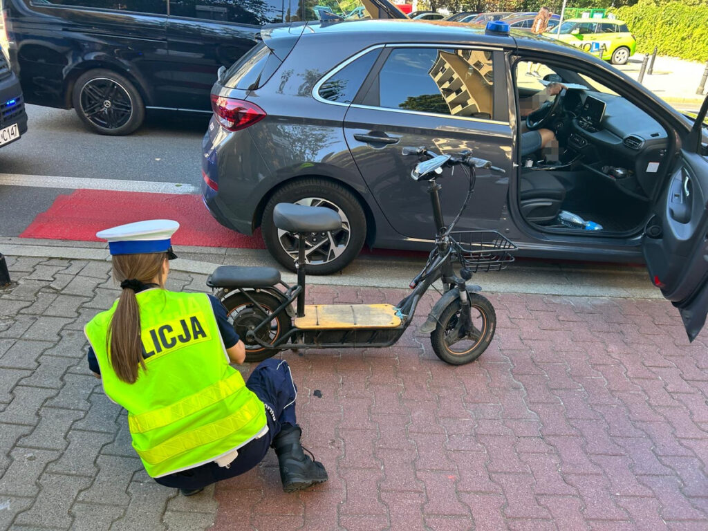 Policjantka w kamizelce z napisem "POLICJA" bada elektryczną hulajnogę na chodniku obok zaparkowanego samochodu.