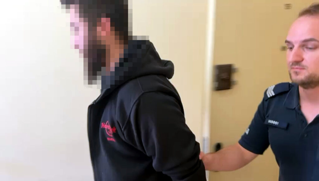 Mężczyzna w czarnej bluzie z kapturem prowadzony przez policjanta w mundurze w korytarzu budynku.