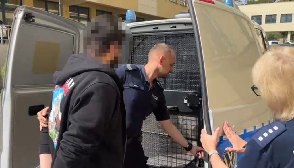 Mężczyzna w ciemnej bluzie wchodzi do policyjnego radiowozu, a policjant otwiera drzwi pojazdu.