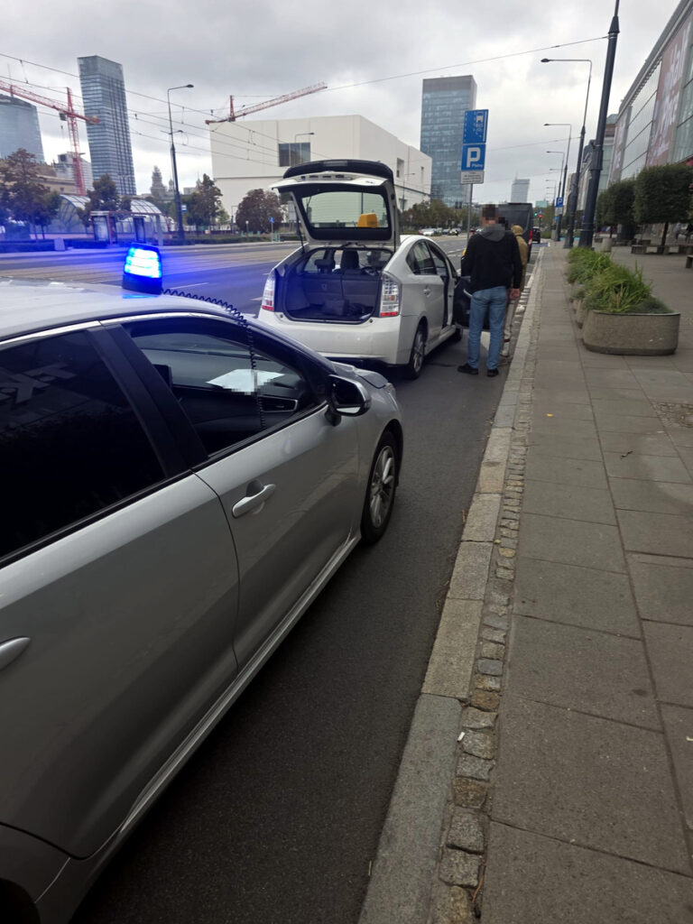 Policyjny radiowóz z włączonym sygnałem świetlnym stoi obok białego samochodu osobowego na ulicy, obok mężczyzny rozmawiającego z kierowcą.