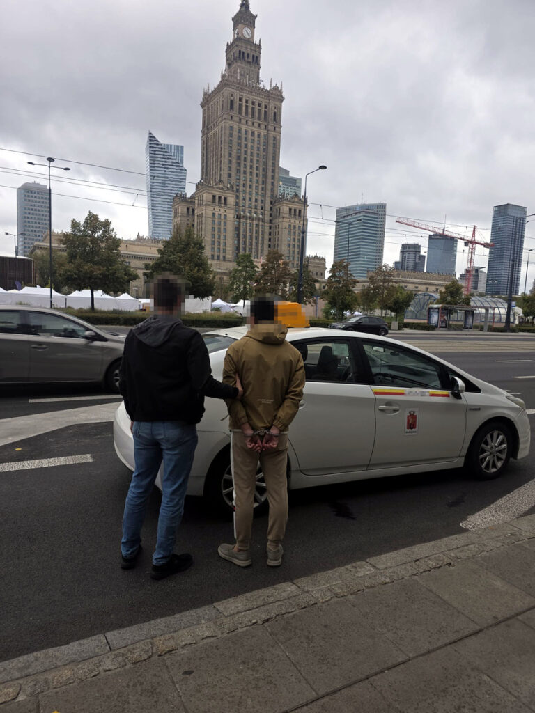 Mężczyzna w czarnej kurtce trzyma innego mężczyznę w kajdankach obok taksówki, w tle widoczny wysoki budynek z zegarem.