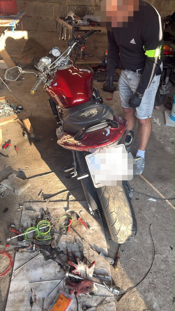 Mężczyzna w rękawicach pracuje przy czerwonym motocyklu w warsztacie, otoczony narzędziami i częściami mechanicznymi.