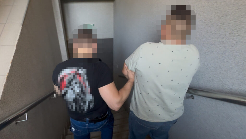Mężczyzna w czarnej koszulce jest prowadzony przez policjanta schodami w budynku.