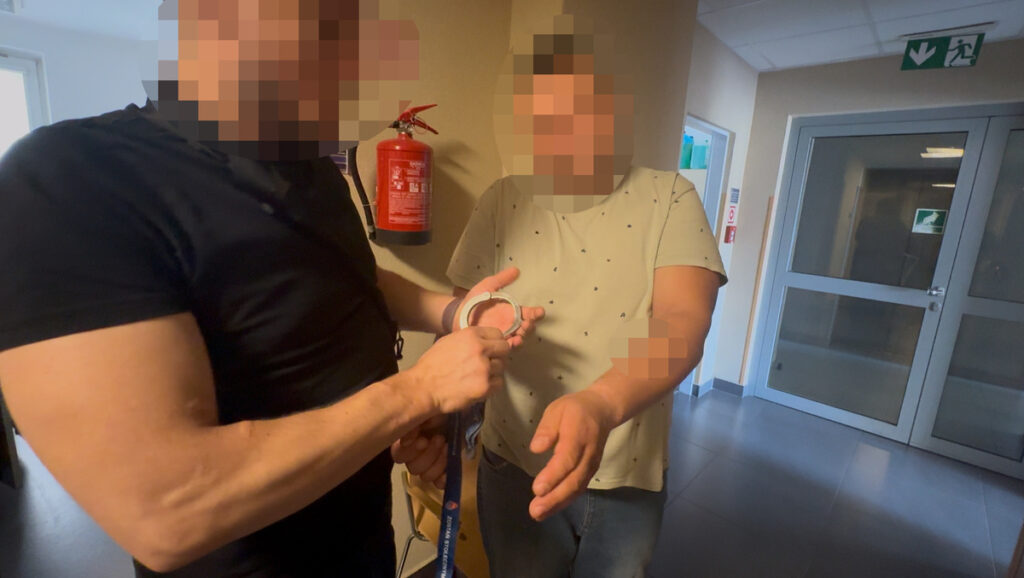 Funkcjonariusz policji zakłada kajdanki mężczyźnie w jasnej koszuli w korytarzu budynku. W tle widoczny gaśnica.