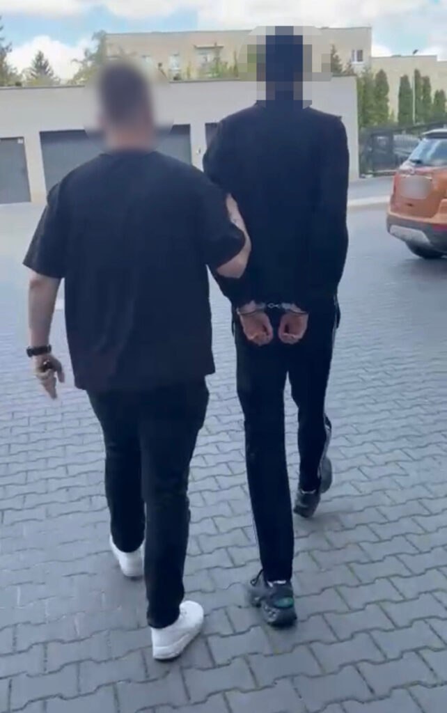 Mężczyzna w czarnej odzieży prowadzony przez drugiego mężczyznę w czarnym t-shircie, obaj na chodniku.