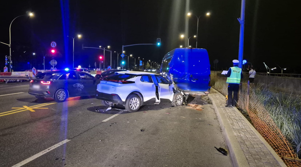 Kolizja drogowa z udziałem białego samochodu osobowego i niebieskiego busa na nocnej ulicy. Policjant dokonuje oględzin.