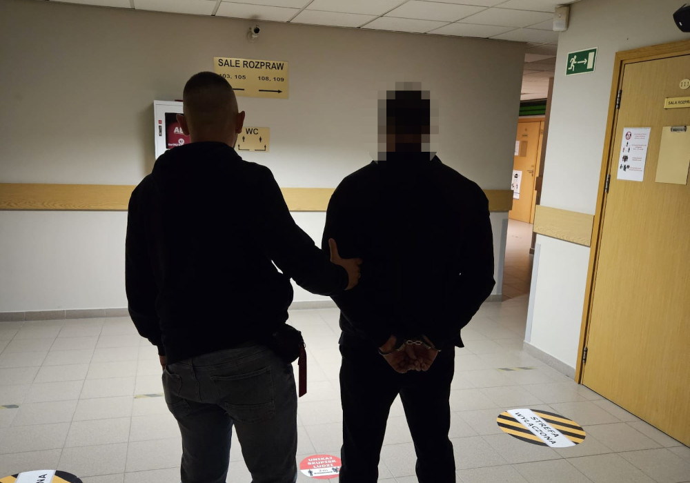Mężczyzna w kajdankach prowadzony przez funkcjonariusza w korytarzu budynku sądu.