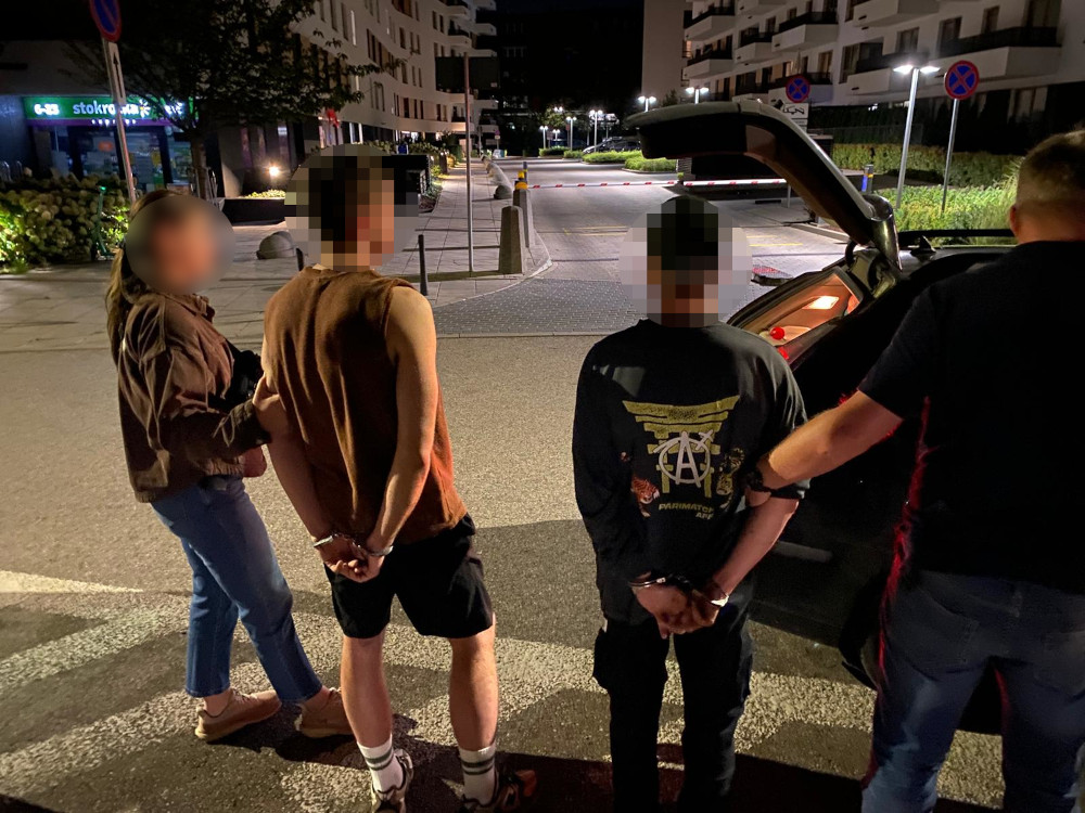 Grupa młodych ludzi stoi obok otwartego bagażnika samochodu, a policjant przeszukuje pojazd w nocnej scenerii.