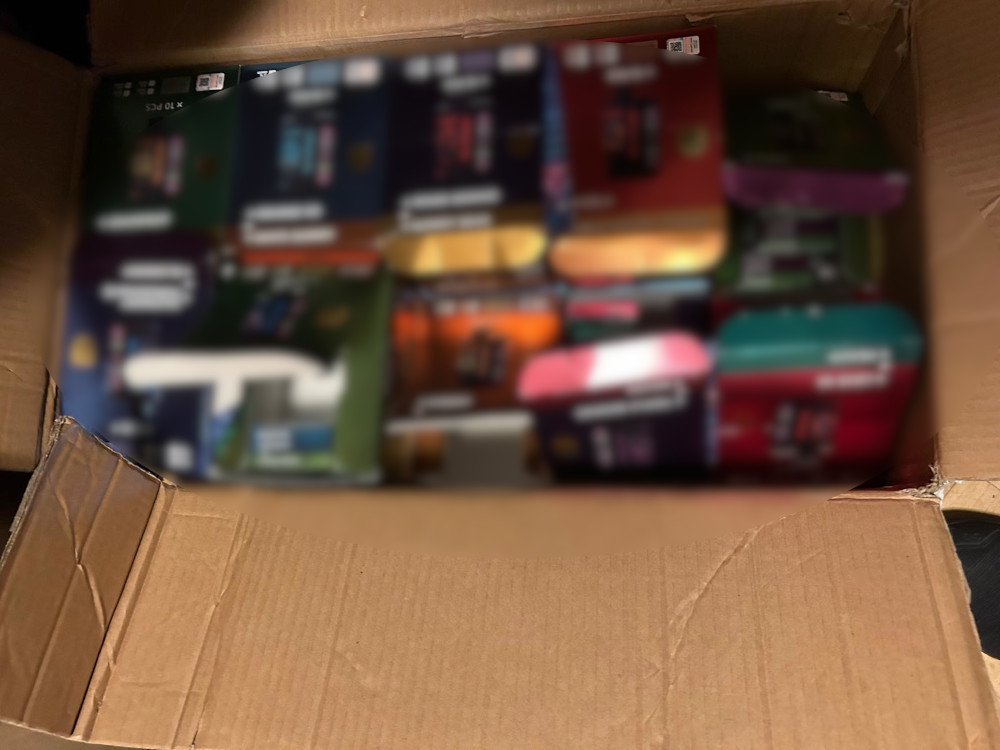Kartonowe pudełko wypełnione kolorowymi opakowaniami różnych produktów, widoczne są tylko ich fragmenty.