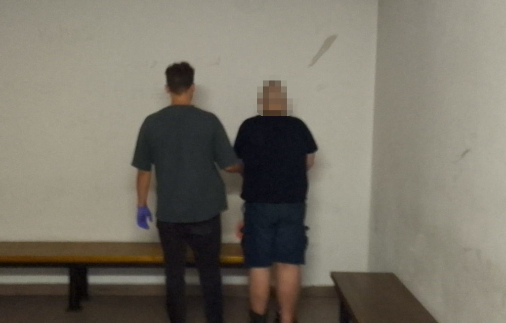 Mężczyzna w rękawiczkach prowadzi osobę w czarnym t-shircie w kierunku ściany w pustym pomieszczeniu.