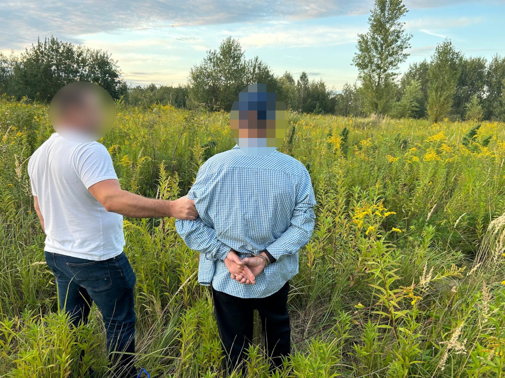 Mężczyzna w koszuli w kratę stoi z rękami za plecami, a inny mężczyzna w białej koszulce trzyma go za ramię w zielonym terenie.
