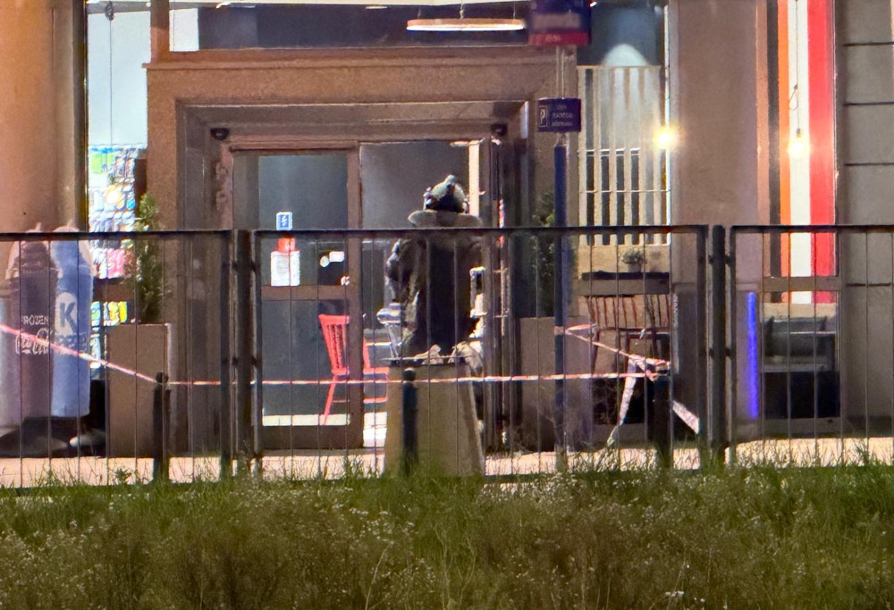 Kontrterrorysta w specjalnym umundurowaniu przeszukuje teren przed wejściem do budynku, otoczony taśmą zabezpieczającą.