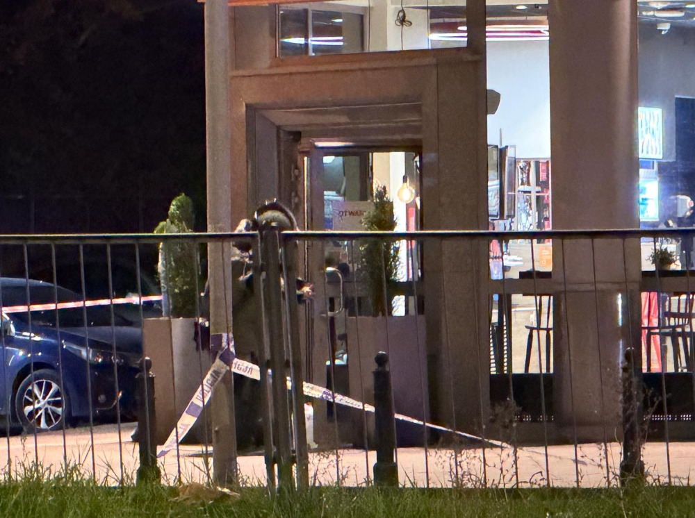 Mężczyzna w kamizelce ochronnej przeszukuje teren przed wejściem do lokalu, zabezpieczonego taśmą policyjną.