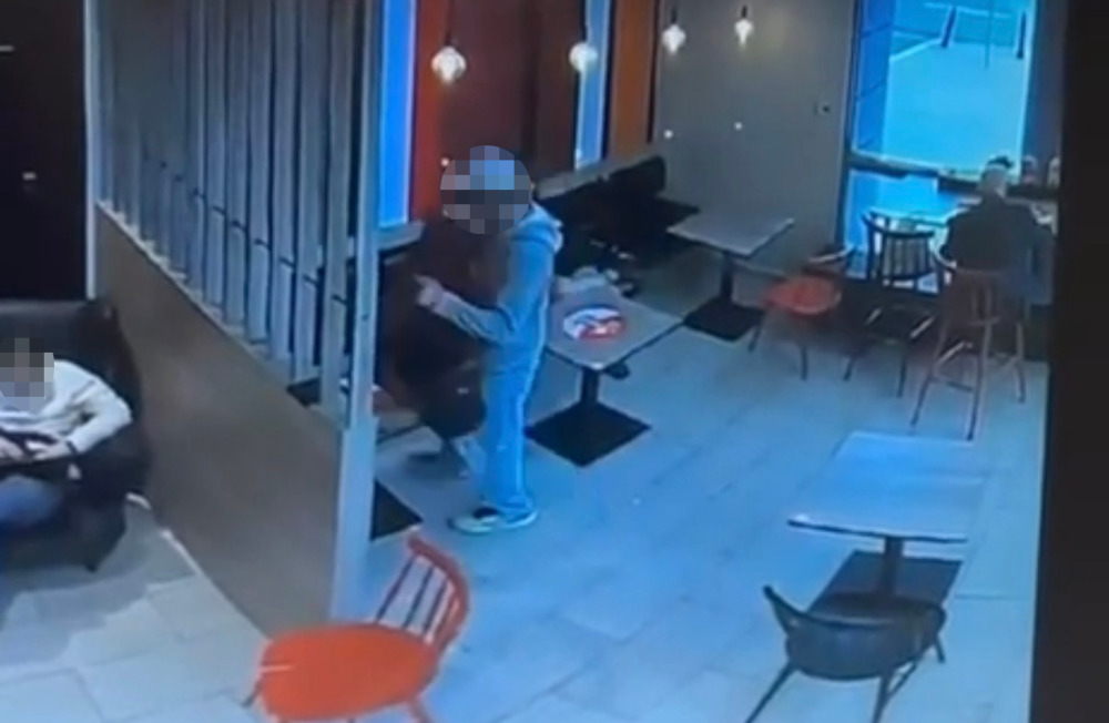 Mężczyzna w szarym dresie rozmawia z osobą przy stole w kawiarni, obok siedzi inny klient na kanapie.
