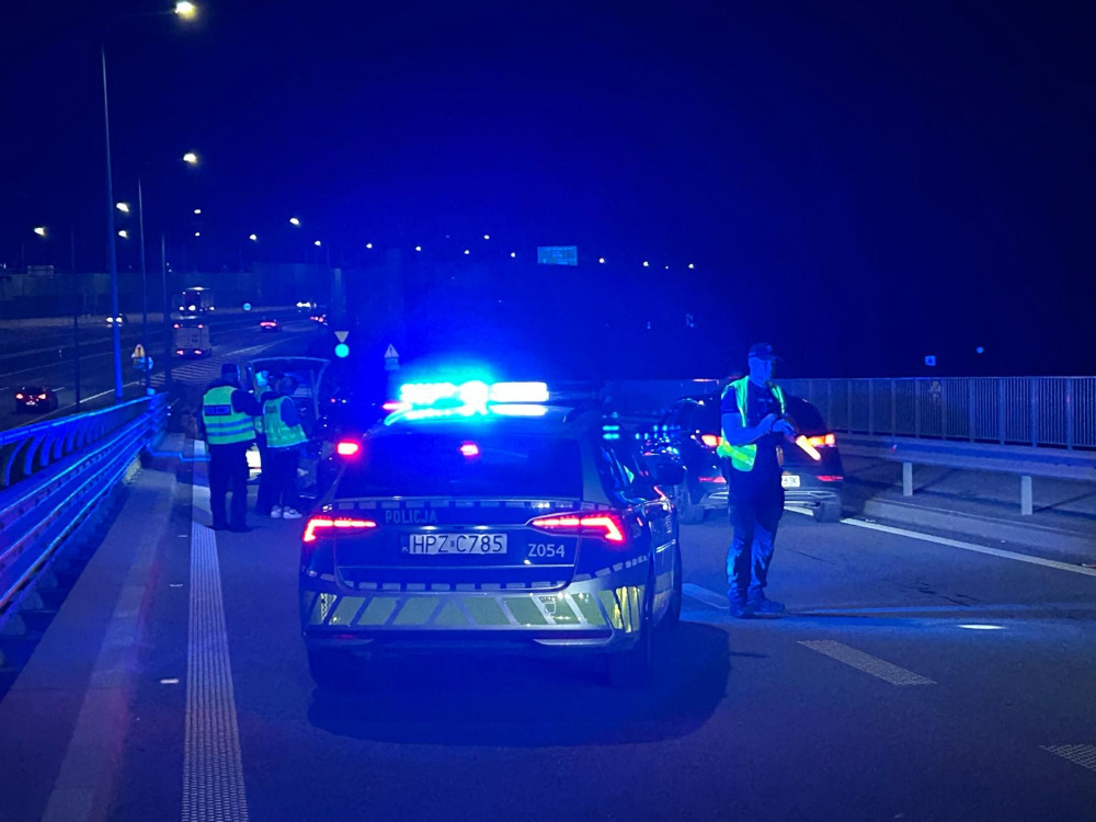 Policjanci w kamizelkach odblaskowych kontrolują pojazdy na nocnej drodze, przy włączonych sygnałach świetlnych.