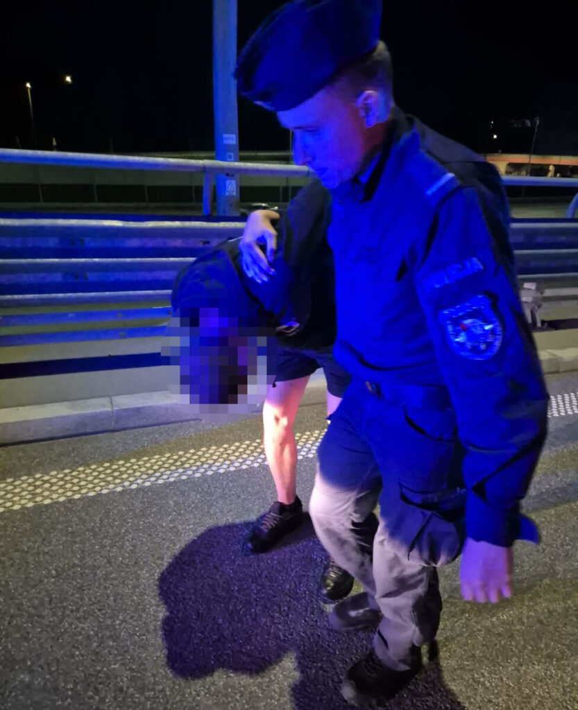Mundurowy funkcjonariusz pomaga poszkodowanemu mężczyźnie na drodze w nocy, oświetlonej przez niebieskie światła.