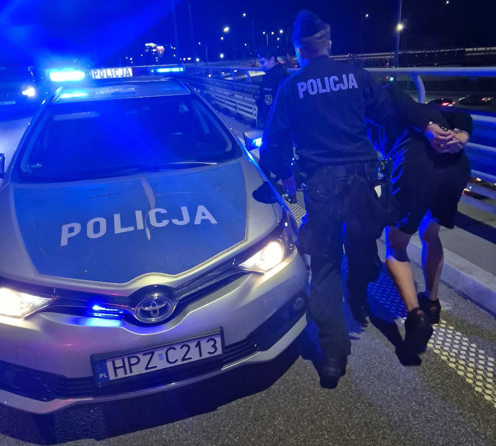 Policjant w mundurze zatrzymuje osobę w okolicy radiowozu policyjnego z włączonymi sygnalizatorami świetlnymi.