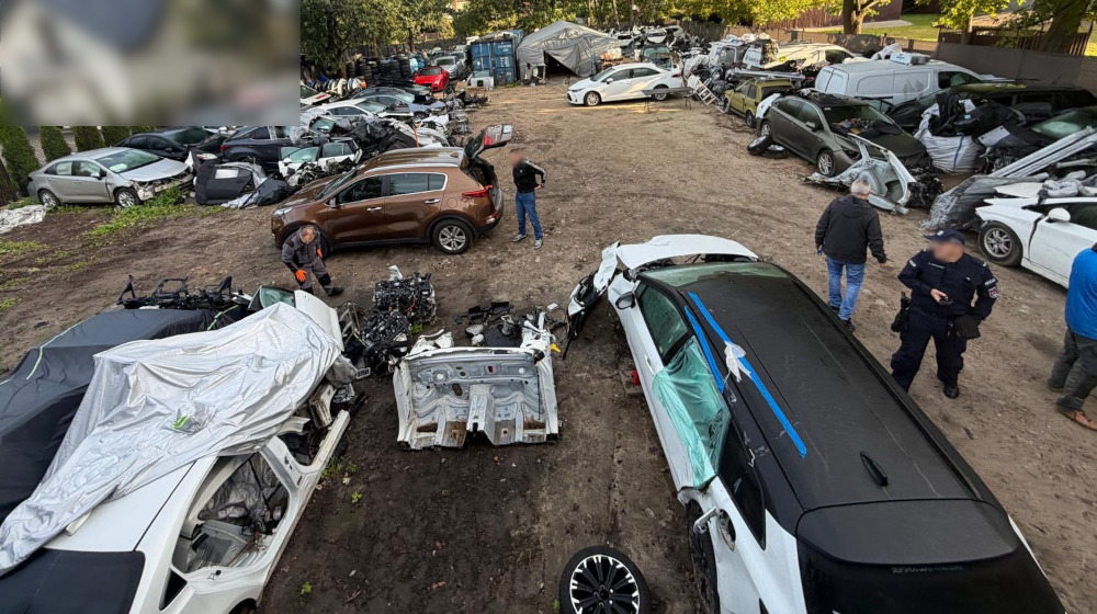 Na terenie złomowiska mężczyzna w roboczym stroju przegląda uszkodzone pojazdy, a obok stoi samochód osobowy.