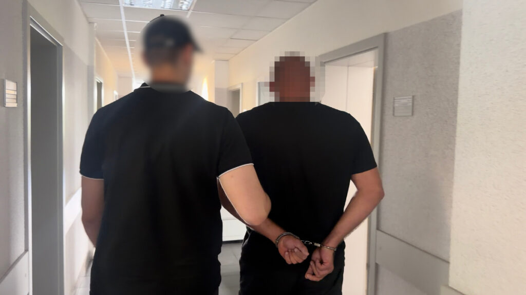 Funkcjonariusz policji prowadzi mężczyznę w kajdankach przez korytarz budynku.