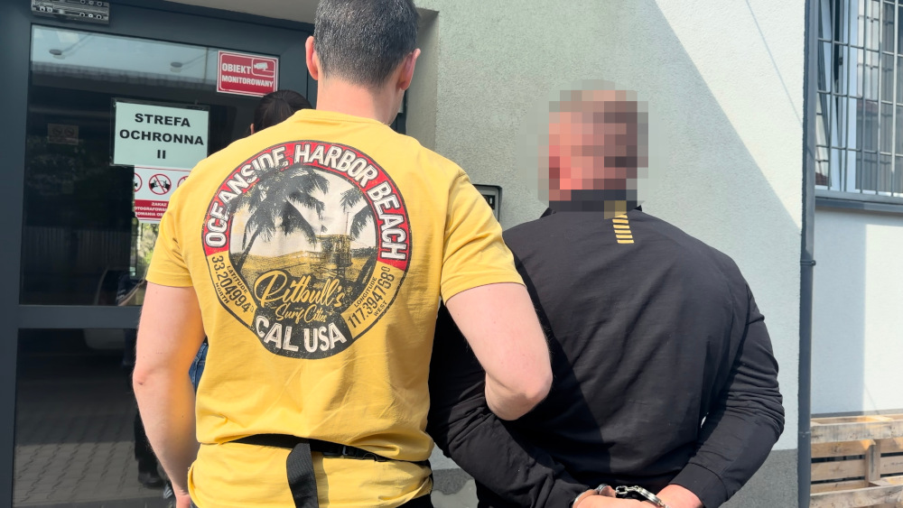Mężczyzna w żółtej koszulce prowadzi osobę w kajdankach w kierunku budynku z oznaczeniem "STREFA OCHRONNA II".