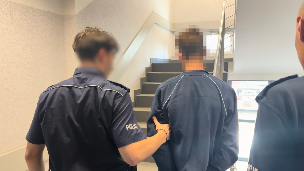 Policjant prowadzi zatrzymanego mężczyznę po schodach w budynku.