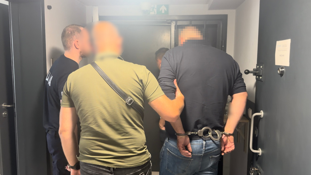 Mężczyzna w kajdankach prowadzony przez funkcjonariusza policji w korytarzu budynku.