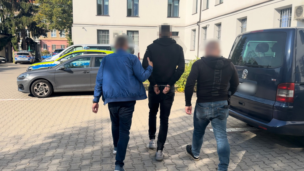 Mężczyzna w kajdankach prowadzony przez dwóch policjantów na parkingu przed budynkiem.