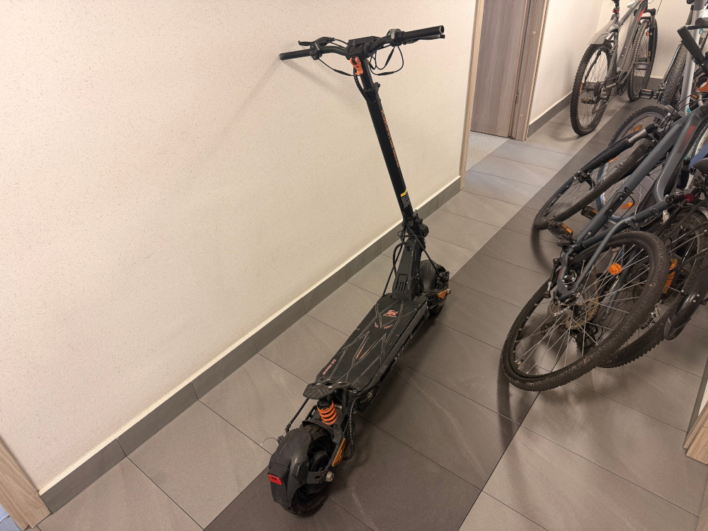 Elektroniczna hulajnoga stojąca w wąskim korytarzu obok zaparkowanych rowerów.