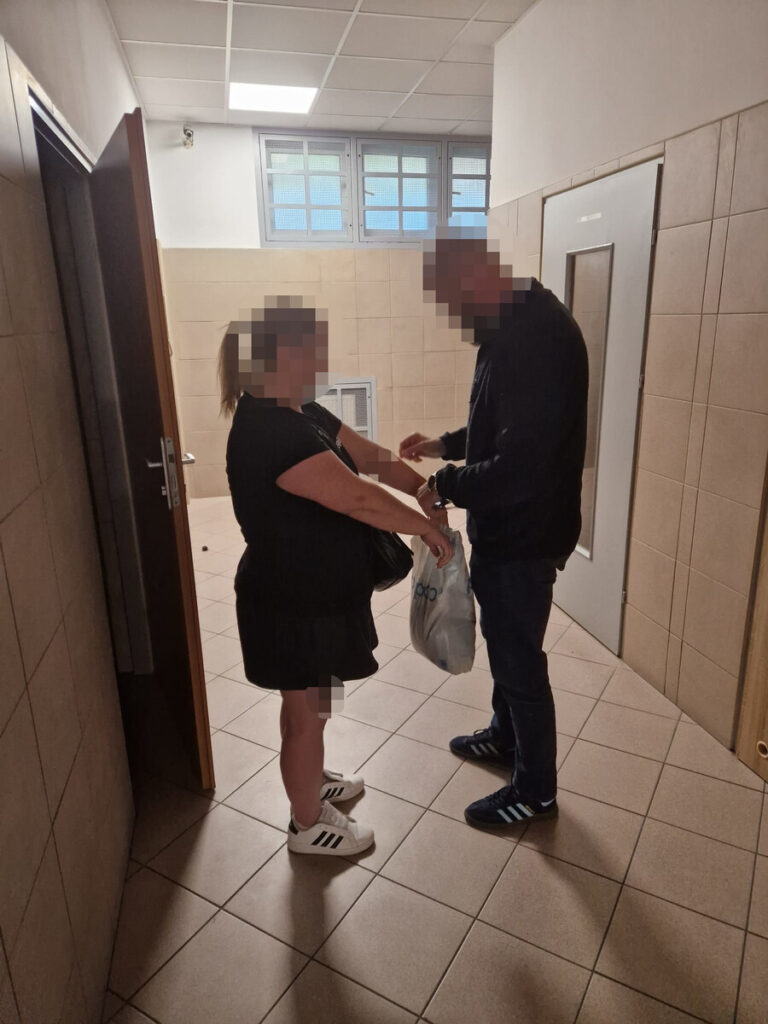 Kobieta i mężczyzna stoją naprzeciw siebie w wyłożonym kafelkami korytarzu; mężczyzna trzyma reklamówkę.
