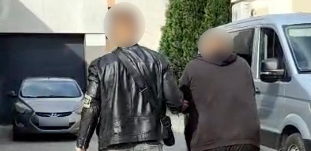 Dwóch mężczyzn z zasłoniętymi twarzami idzie między zaparkowanymi samochodami przy budynku.