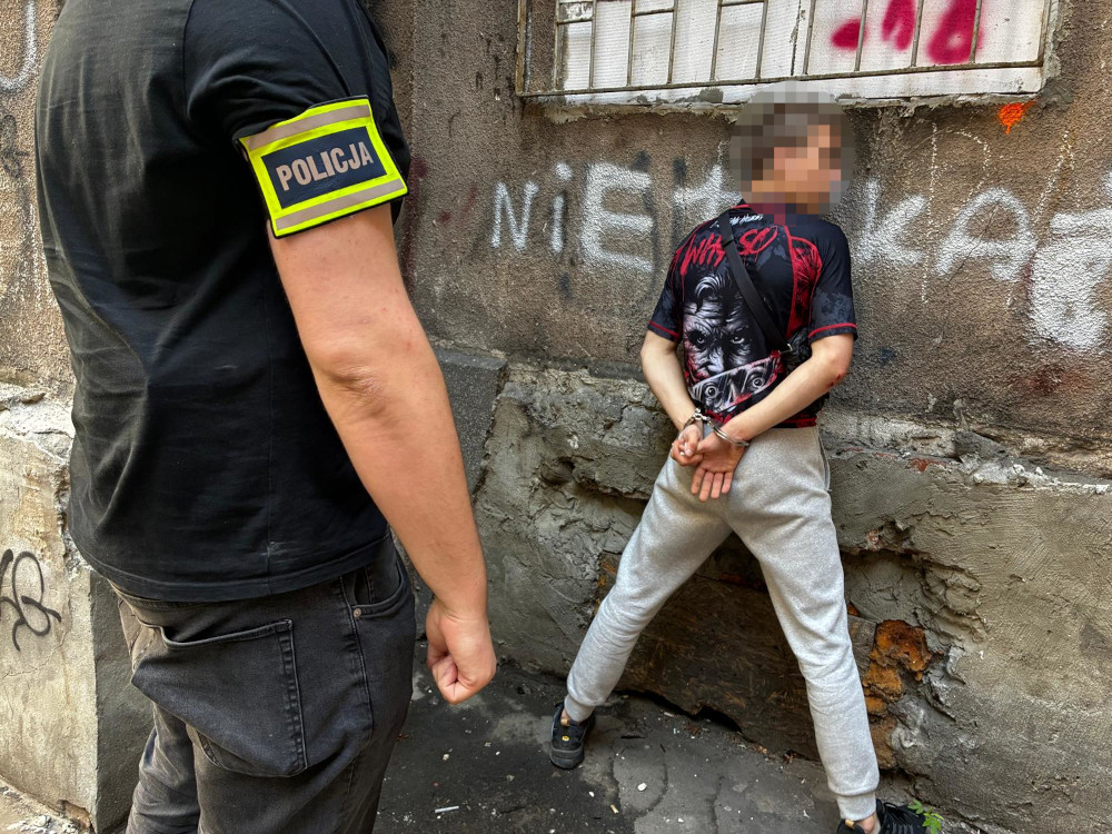 Policjant w koszulce z napisem "POLICJA" zatrzymuje młodego mężczyznę w szarych spodniach, przy ścianie z graffiti.