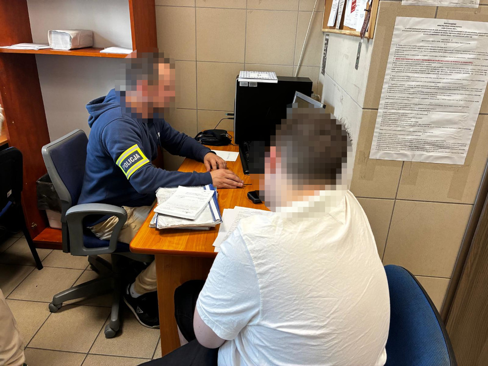 Policjant w mundurze rozmawia z młodym mężczyzną siedzącym przy biurku w pomieszczeniu biurowym.