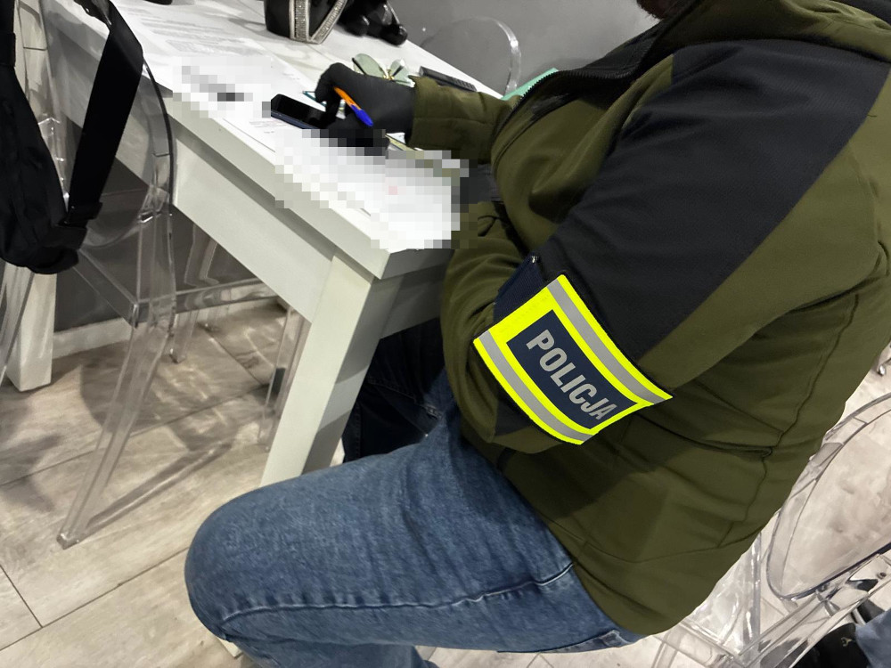 Funkcjonariusz policji w zielonej kurtce z oznakowaniem "POLICJA" siedzi przy stole, pisząc na kartce.